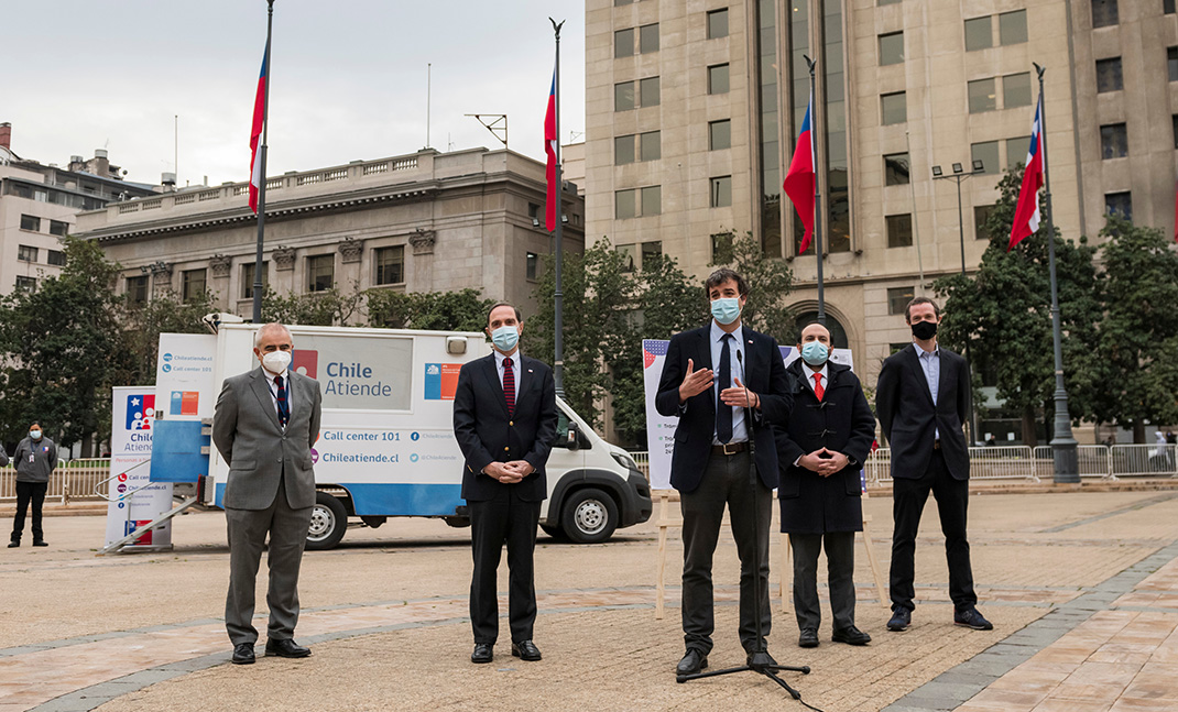 Autoridades de Gobierno en la Plaza de la Constitución, atrás el móvil del IPS y banderas chilenas en sus mástiles.